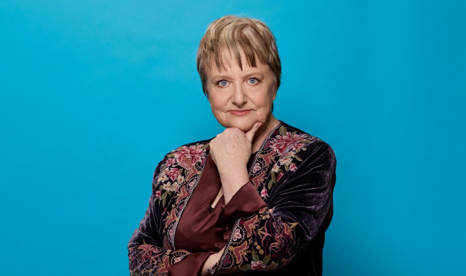 Denise Scott poses in front of a blue background. She wears a floral embroidered velvet jacket.