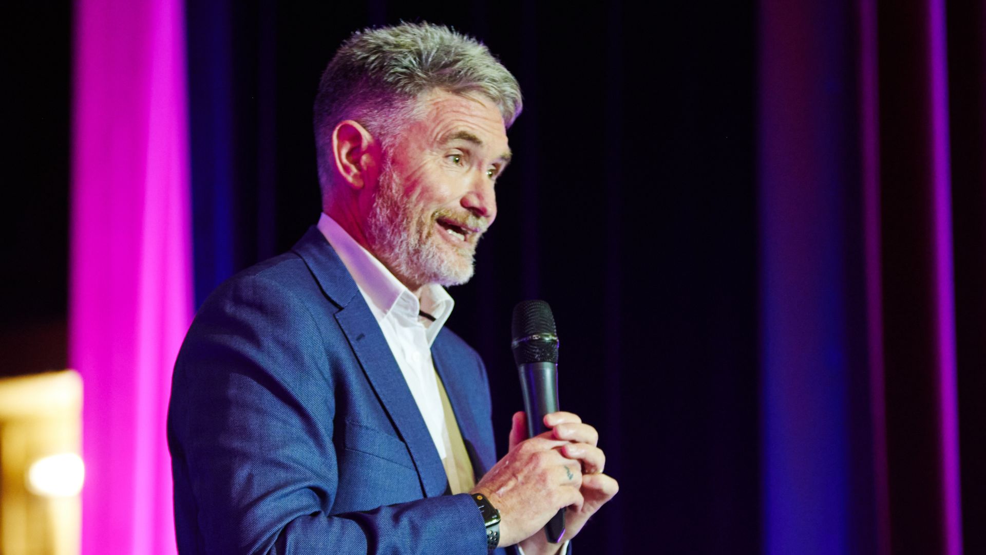 Dave Hughes as MC at a corporate function. He wears a blue suit and holds a microphone.